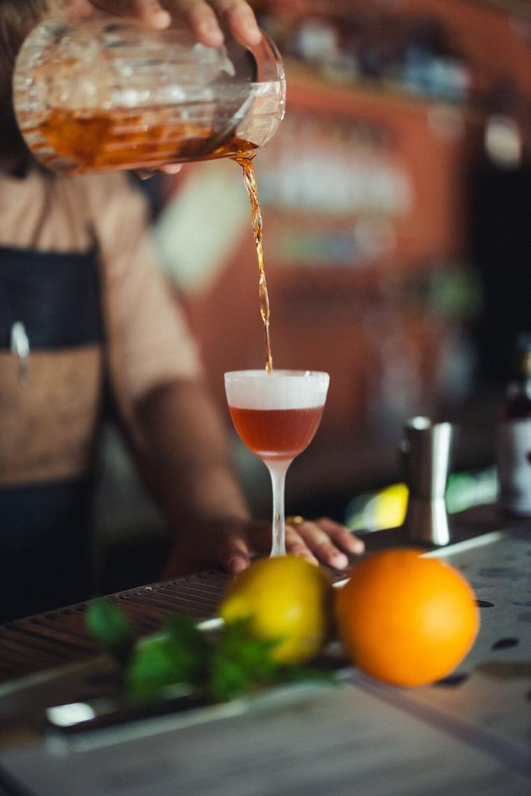 Rødlig cocktail hældes fra røreglas i en coupe med citron og appelsin ved siden af